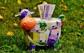 Копилка корова с купюрами евро на зеленой траве 