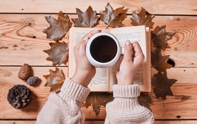 Cup of coffee in hands on a table with a book and leaves