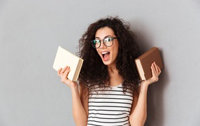 Веселая девушка в очках с книгами на фоне серой стены 