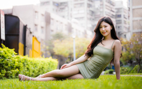 Улыбающаяся азиатка в платье сидит на зеленой траве 