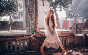 Молодая девушка азиатка в белом платье сидит на разваленной стене