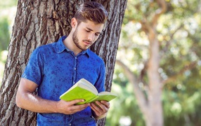 Handsome young man stands with a book under a tree