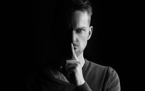 Молодой мужчина закрыл рот пальцем на черном фоне 