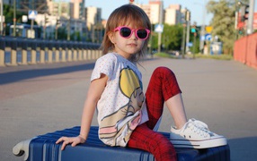 Маленькая девочка в очках сидит на чемодане
