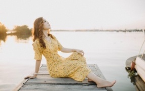 Красивая девушка в платье сидит на мосту у реки