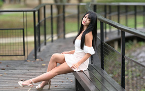Beautiful long-haired Asian woman in a white dress sits on a bench