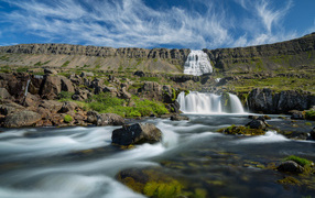 A waterfall flows down from the mountains against the backdrop of the beautiful blue sky of Iceland