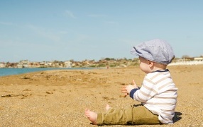 A little boy in a cap sits on the sand near the water