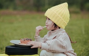 Маленькая девочка в желтой шапке ест пирожное