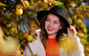 Улыбающаяся девушка в черной шляпе у дерева с яблоками