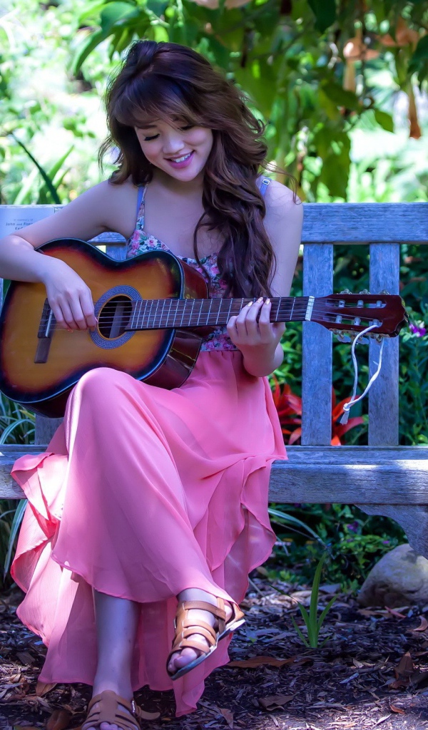 Девушка сидит на скамье в саду и играет на гитаре