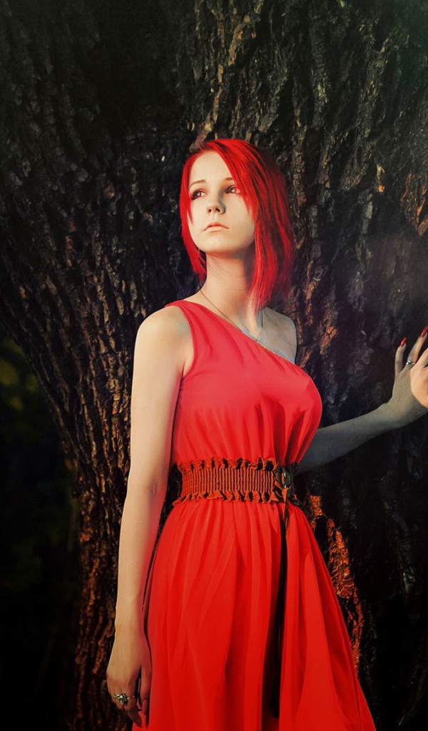 Рыжая девушка в красном платье стоит у дерева