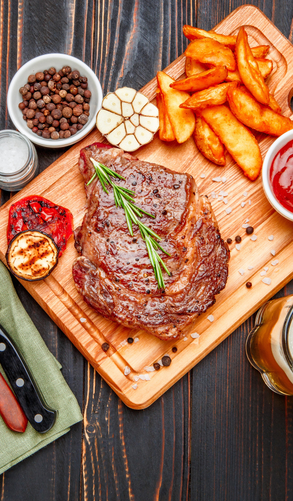 Сочный кусок мяса с картофелем фри, специями и бокалом пива на толе