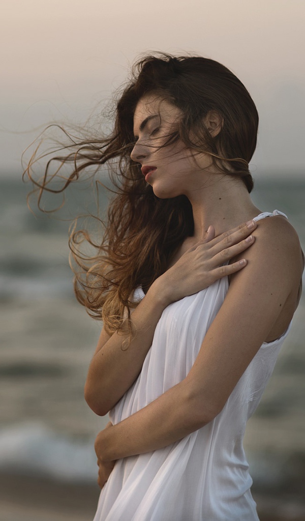 Мечтательная девушка в белом наряде стоит на берегу океана