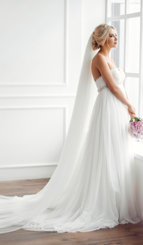 Красивая девушка в белом свадебном платье с букетом стоит у окна