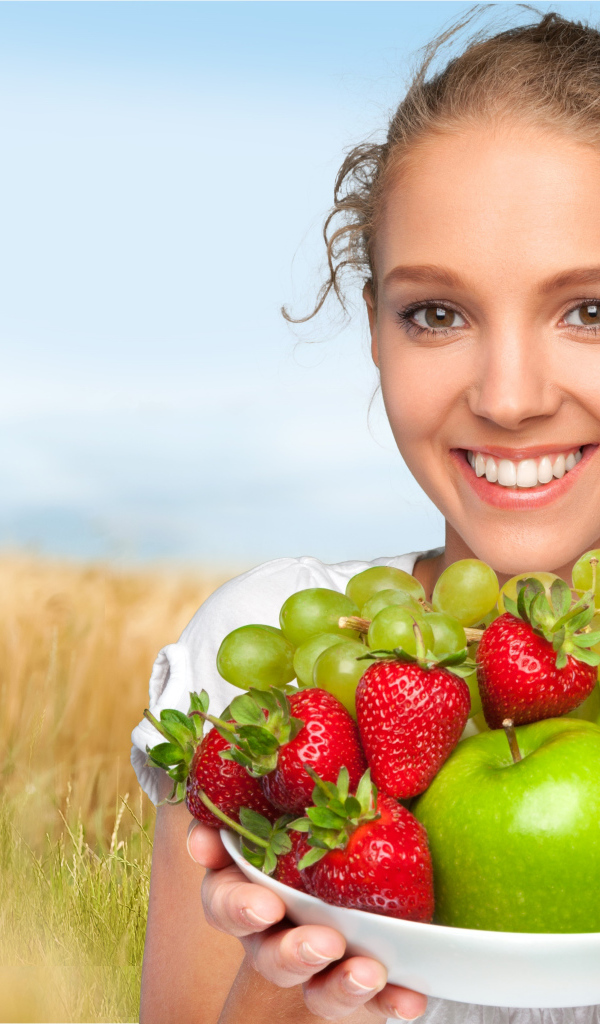 Улыбающаяся девушка шатенка с тарелкой с яблоками, виноградом, клубникой и грушей в руках