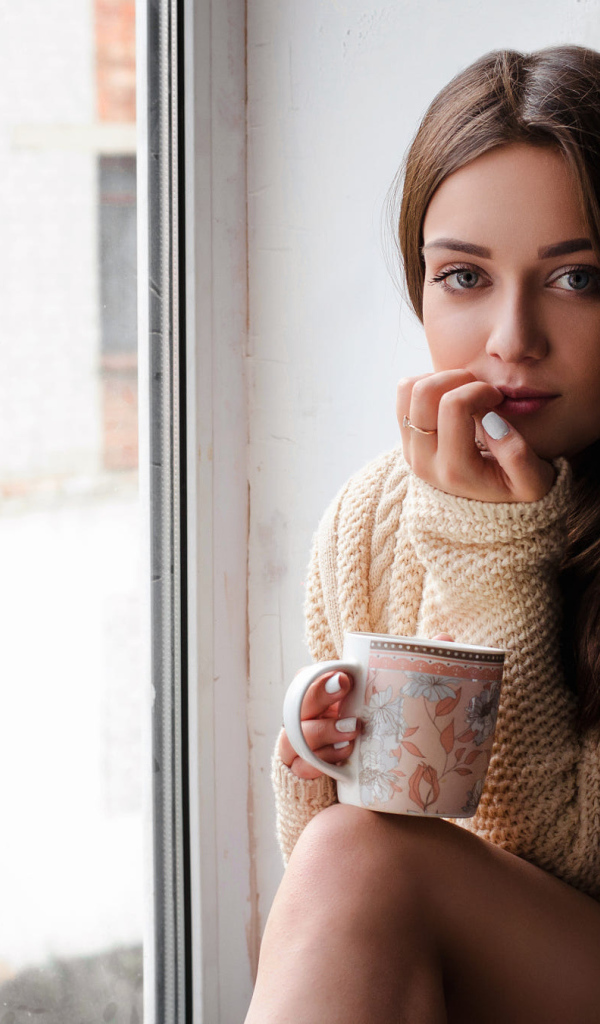 Молодая девушка в свитере сидит с чашкой на подоконнике