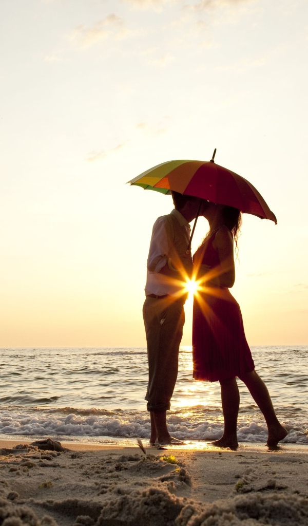 Влюбленная пара на пляже стоит под зонтом на закате