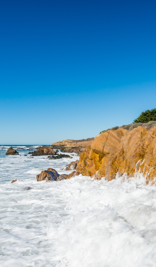 Белые волны бьются о скалы на берегу океана под голубым небом