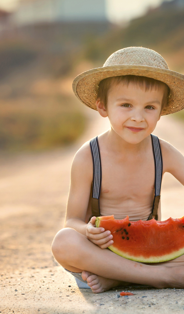 Маленький мальчик в шляпе сидит на дороге с долькой арбуза