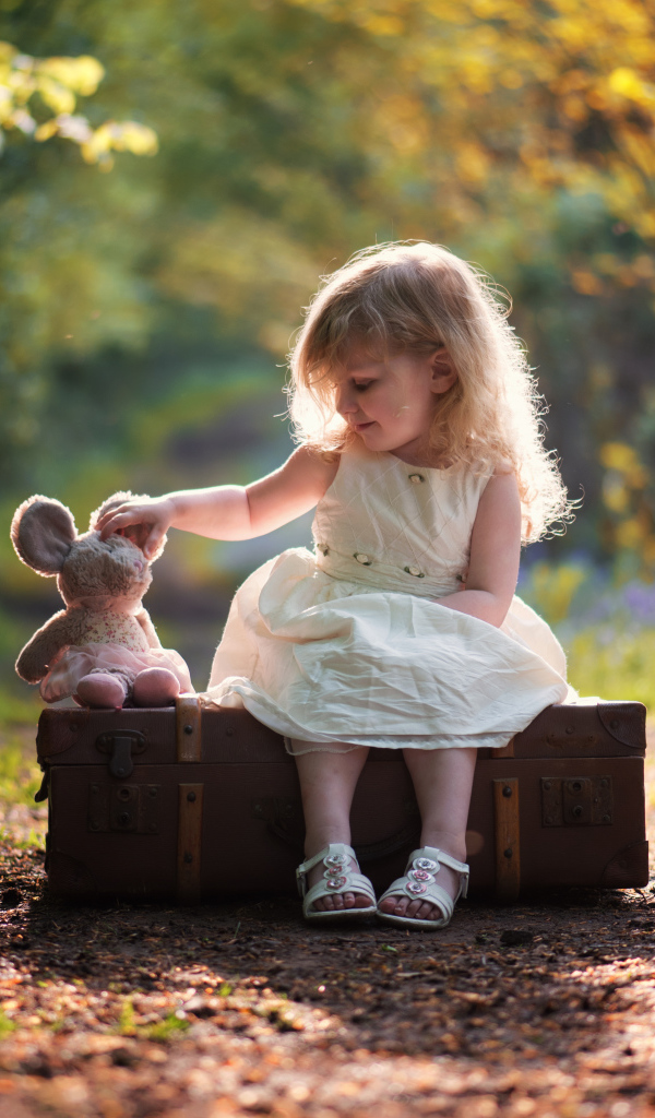 Маленькая девочка в белом платье сидит на большом чемодане с игрушкой