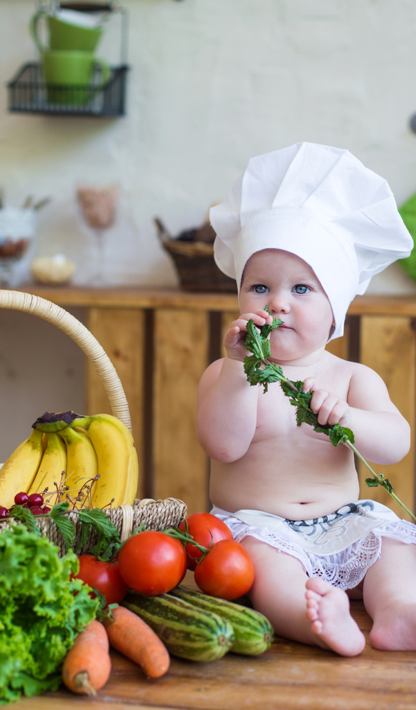 Маленький ребенок в шапке повара сидит на столе с овощами