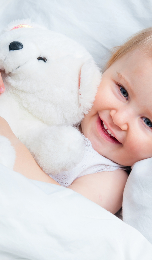 Улыбающийся ребенок лежит в кровати с белым плюшевым медвежонком