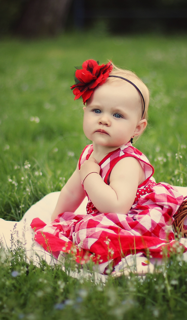 Красивая маленькая девочка с красным цветком на голове сидит на зеленой траве