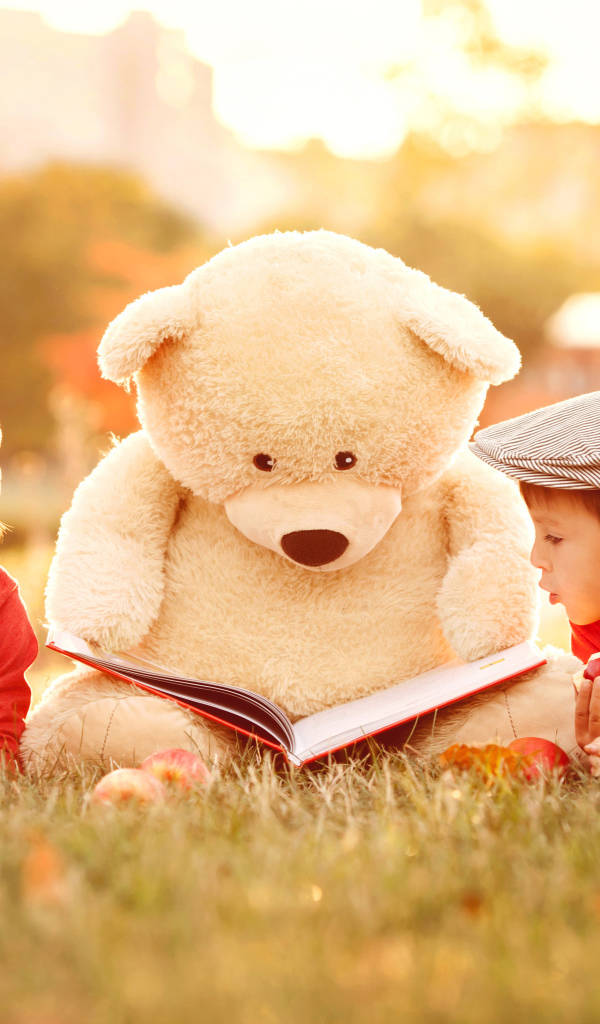 Большой плюшевый медведь с книгой и маленькие мальчики лежат на зеленой траве 