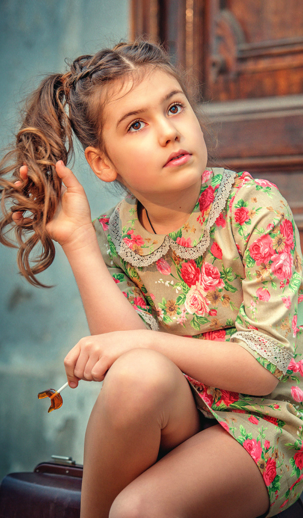 Кареглазая девочка в цветастом платье сидит на пороге