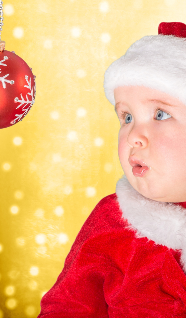 Маленький грудной ребенок в костюме Санты смотрит на елочный шар