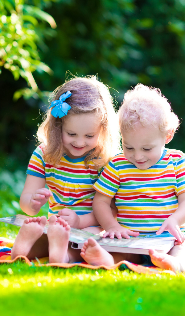 Маленькие мальчик и девочка читают книгу сидя на зеленой траве