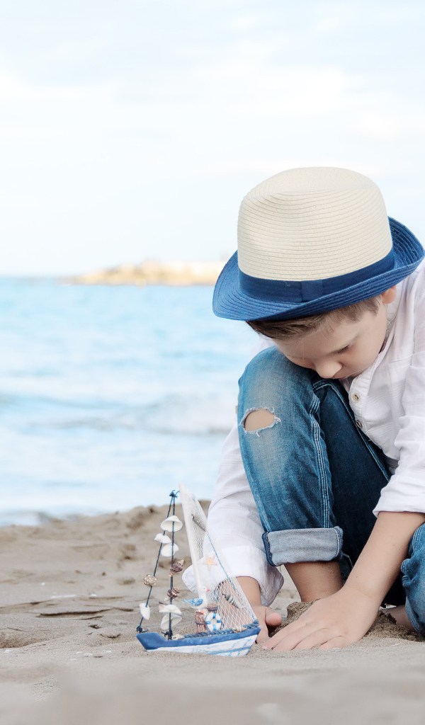 Маленький мальчик в шляпе играет с игрушечным корабликом на песке
