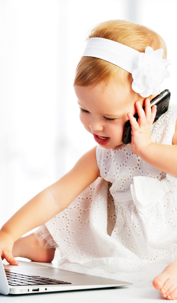Маленькая девочка в белом платье сидит с телефоном у ноутбука