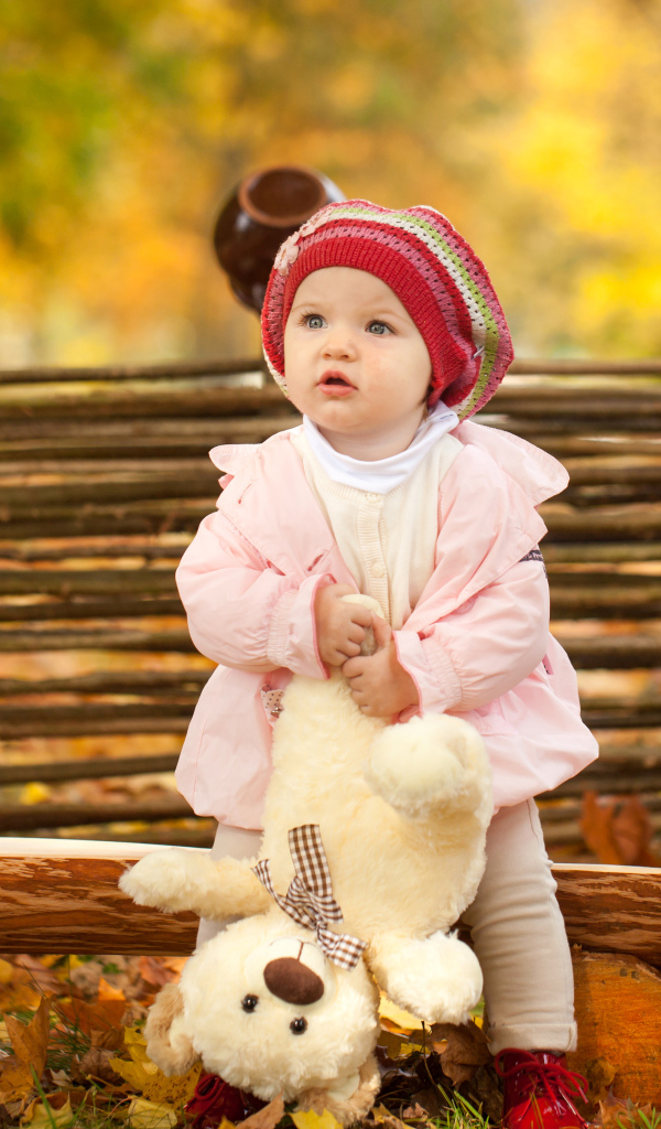 Маленькая девочка с плюшевым мишкой сидит на деревянной лавке осенью