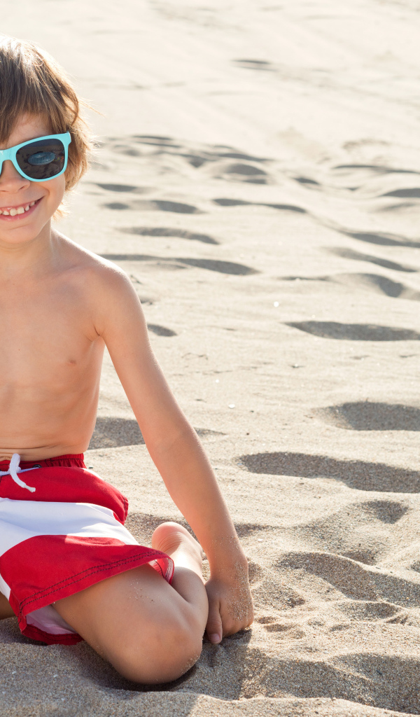 Маленький улыбающийся мальчик в очках сидит на песке