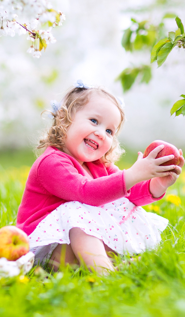 Маленькая улыбающаяся девочка сидит на зеленой траве с яблоками