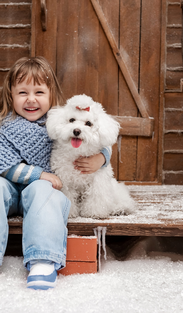 Маленькая улыбающаяся девочка сидит с белым пуделем на крыльце дома зимой