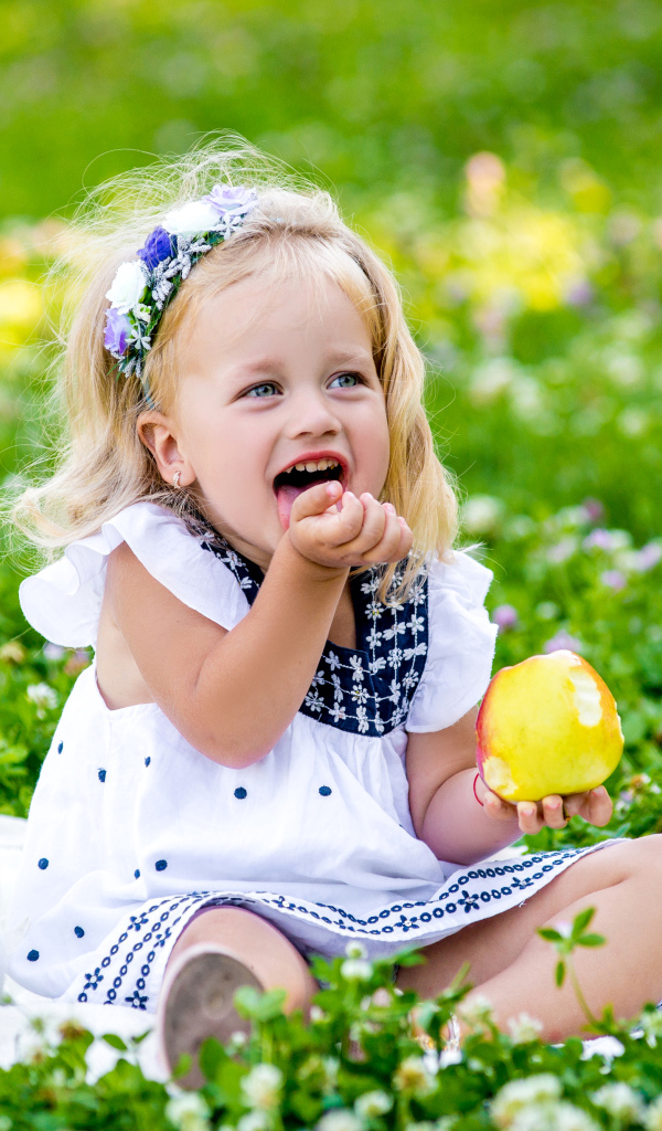 Маленькая улыбающаяся девочка с яблоком сидит на зеленой траве