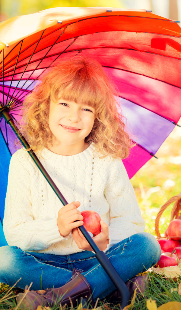 Улыбающаяся девочка с корзиной яблок сидит под разноцветным зонтом