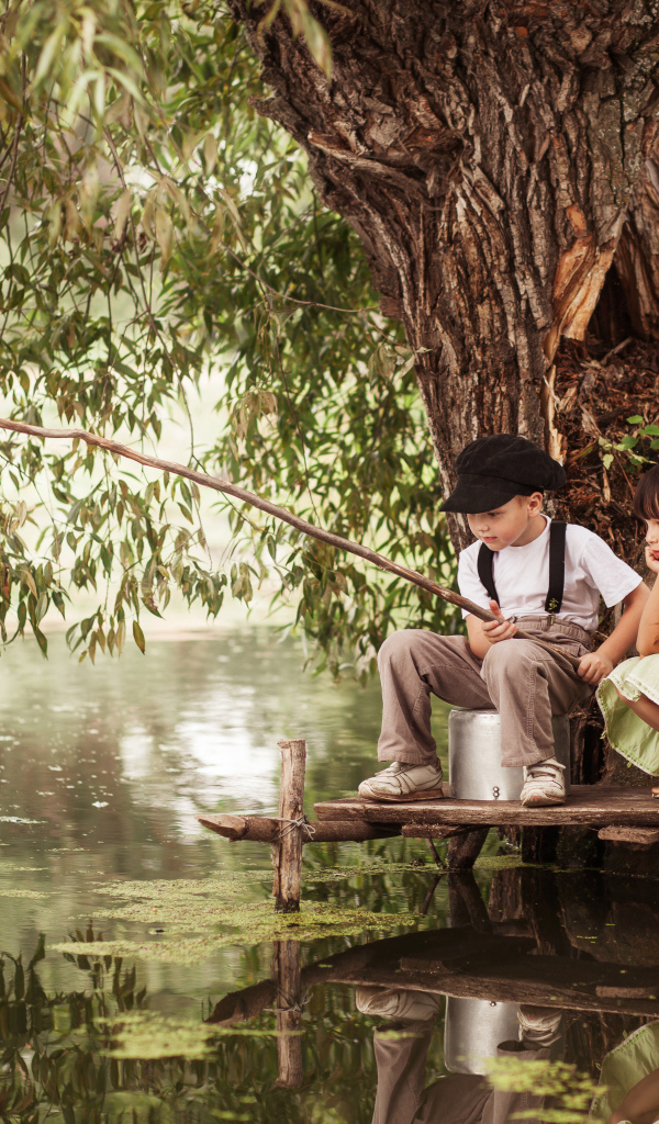 Две маленькие девочки и мальчик сидят на берегу пруда с удочкой