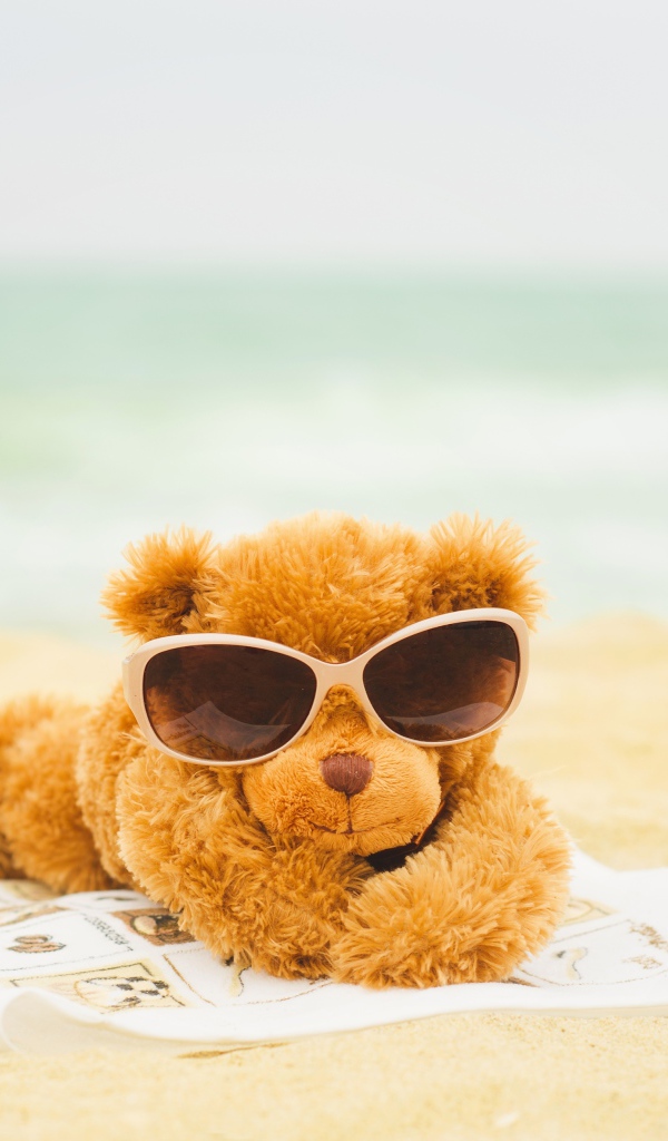 Плюшевый мишка в солнечных очках лежит на пляже