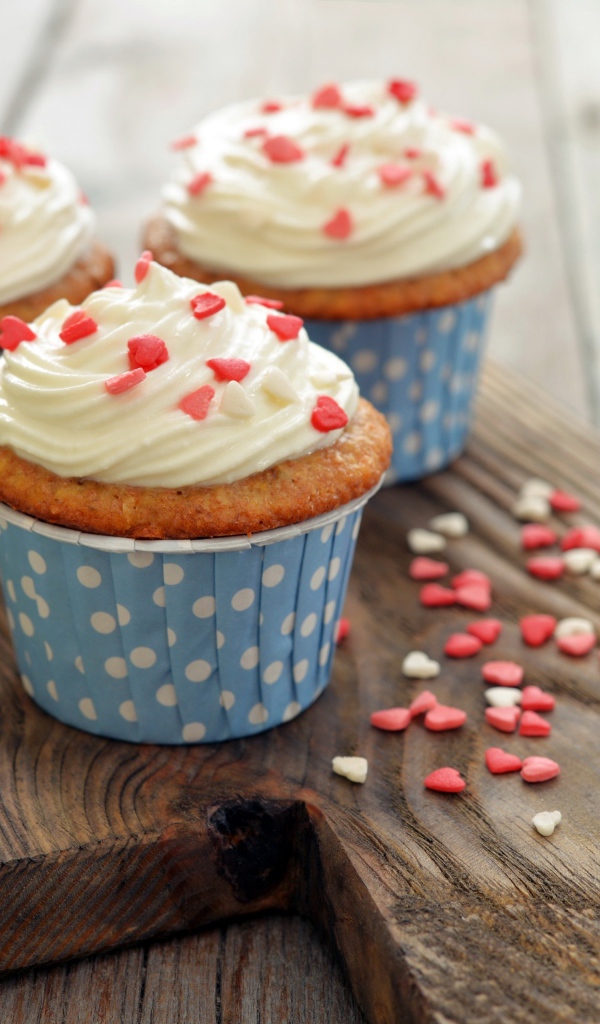 Три кекса со сливками украшены сердечками стоят на разделочной доске