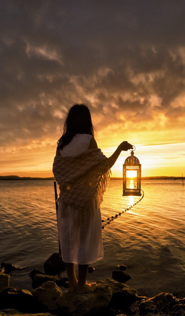Девушка в белом платье стоит у воды с фонарем