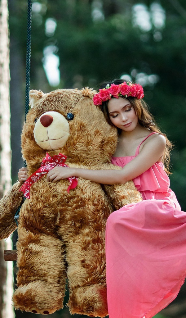 Красивая девушка азиатка в розовом платье сидит на качели с большим игрушечным медведем