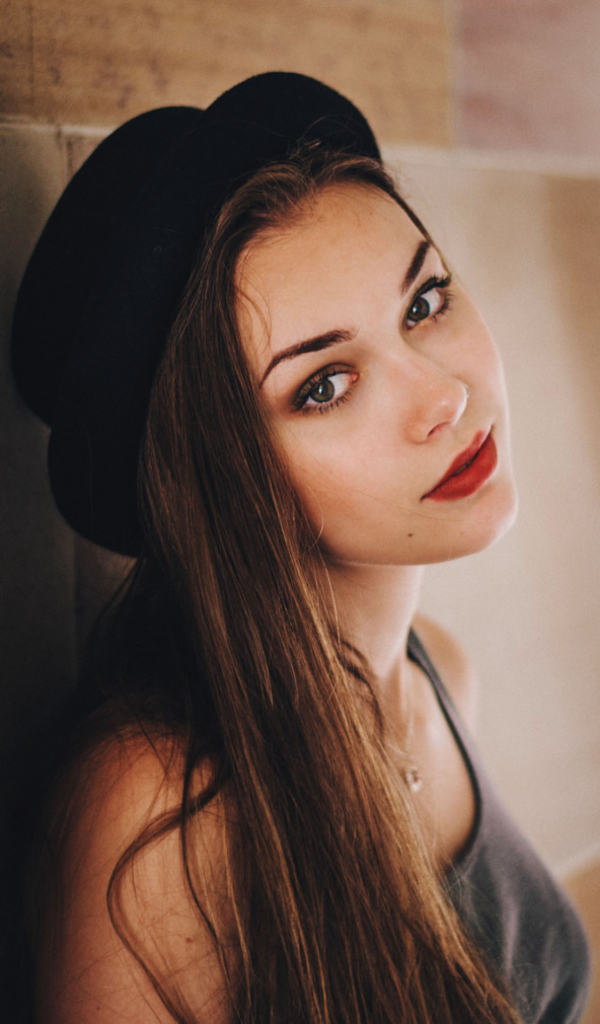 Красивая девушка в черной шляпе у стены 