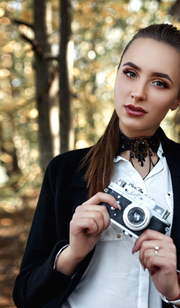 Красивая девушка с фотоаппаратом в руках стоит у дерева