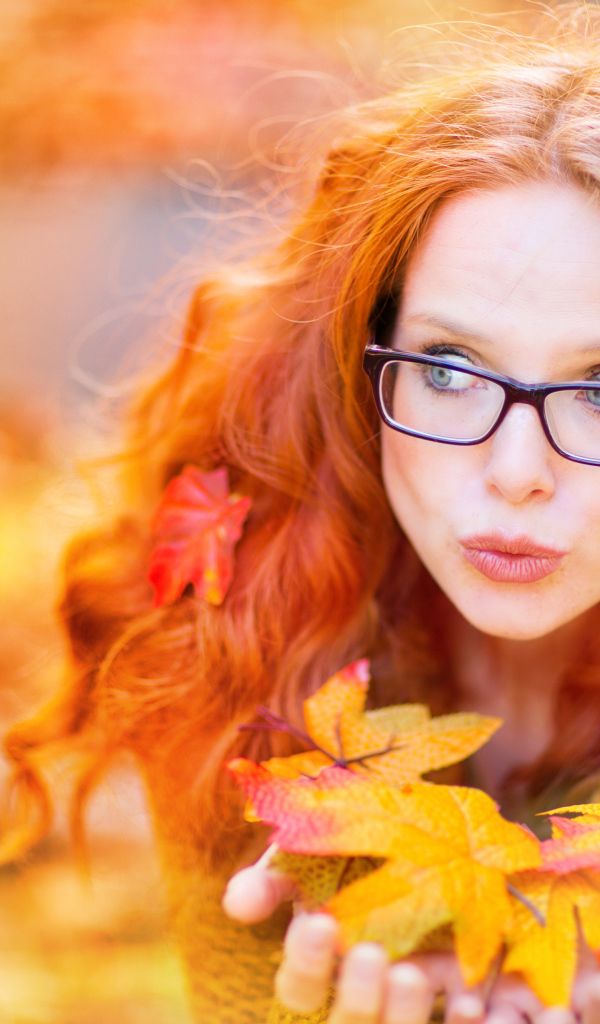Рыжеволосая девушка в очках держит в руках опавшие листья 