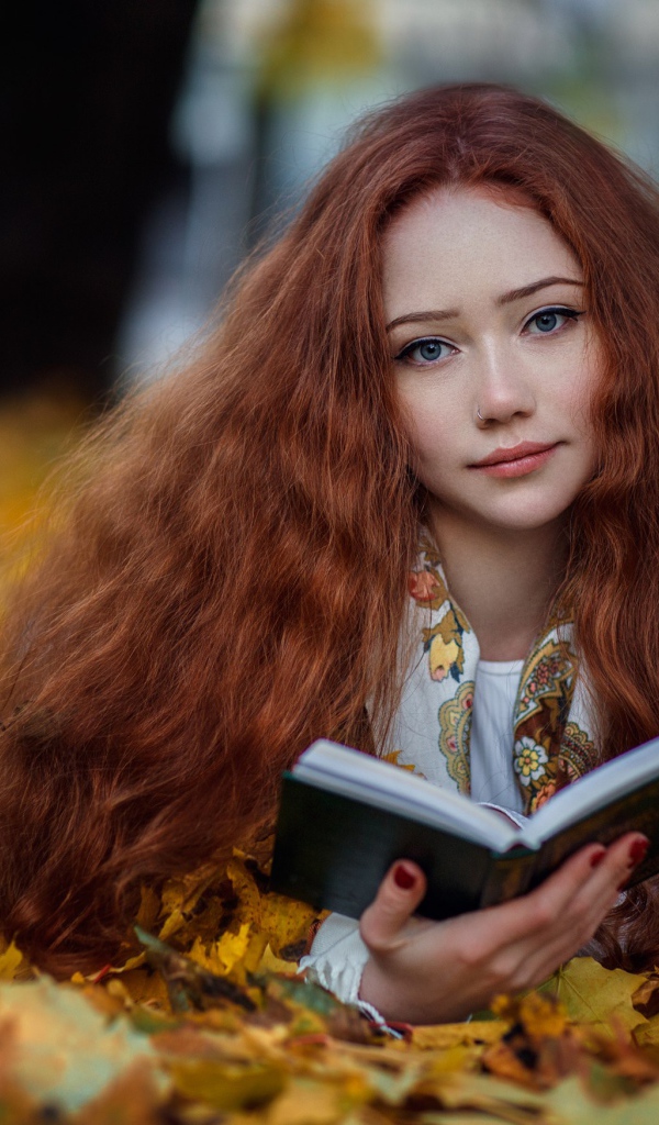 Рыжеволосая девушка с книгой лежит на опавших листьях осенью 