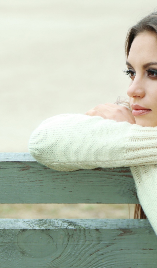Задумчивая девушка брюнетка в белом свитере сидит на лавке